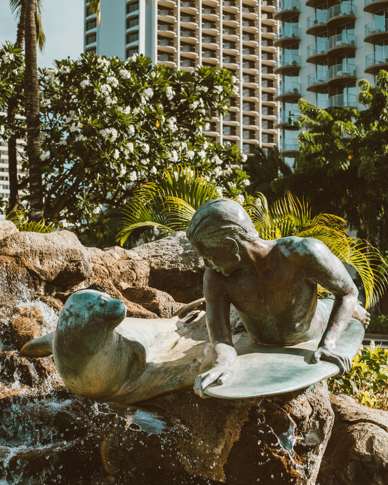 Bronze surfer statue at Kuhio Beach, surrounded by lush greenery and buildings in Waikiki, Oahu.